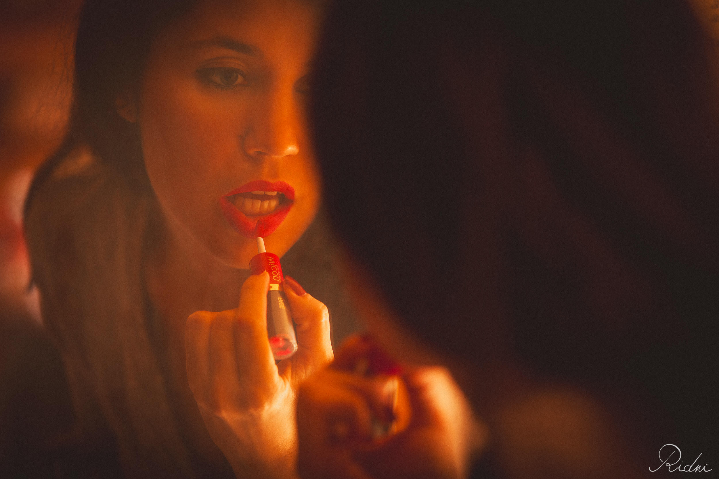 Una donna applica un rossetto rosso vibrante guardandosi allo specchio, con una luce calda e cinematografica a fuoco morbido.