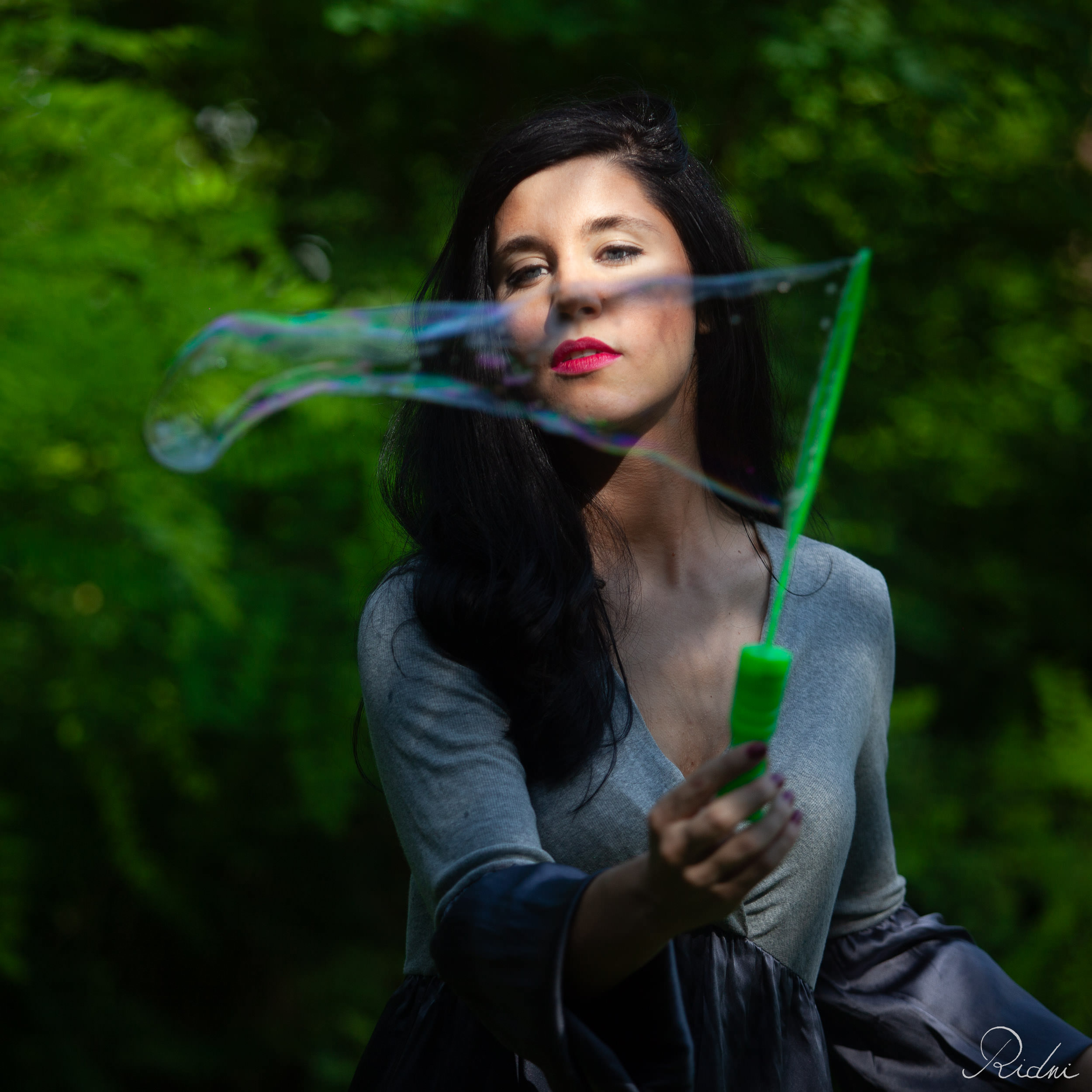 Donna dai capelli scuri con rossetto rosso crea una grande bolla di sapone iridescente in un bosco rigoglioso colpito da luci e ombre.